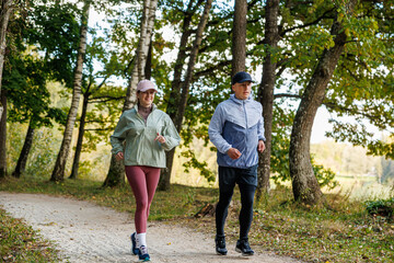 Smiling man and woman jog side by side on a dirt trail lined with trees in early autumn, wearing sporty clothes. Natural background with room for copy space