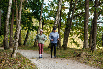 Fototapeta premium Smiling man and woman jog side by side on a dirt trail lined with trees in early autumn, wearing sporty clothes. Natural background with room for copy space