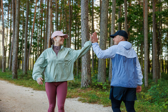 Athletic man and woman share a high five in a pine forest, smiling after exercising. Both wear sporty clothing on a dirt trail. Natural lighting and forest copy space - Powered by Adobe