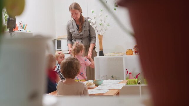 Coach carrying plastic plate while child approaches as she walks to table and drops it adjusting bowl during kindergarten mealtime activity with children seated showing guidance learning