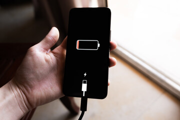 A black smartphone with a low battery icon is connected to a charger on a wooden surface. The charger is plugged into a power outlet.