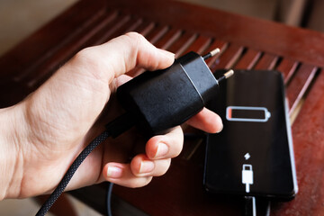 A black smartphone with a low battery icon is connected to a charger on a wooden surface. The charger is plugged into a power outlet.