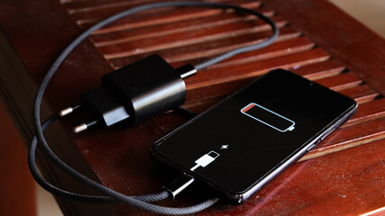 A black smartphone with a low battery icon is connected to a charger on a wooden surface. The charger is plugged into a power outlet.
