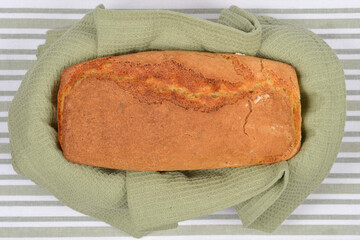 A golden-brown loaf of bread  containing cornmeal, is nestled in a wrinkled sage green waffle-weave towel on a striped background