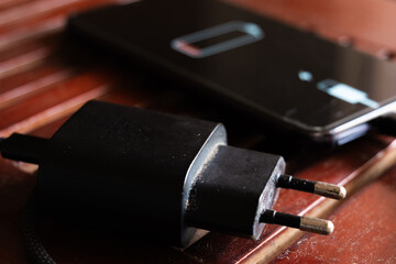 A black smartphone with a low battery icon is connected to a charger on a wooden surface. The charger is plugged into a power outlet.