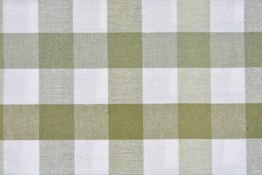A full-frame, close-up shot captures the classic square pattern of a gingham fabric in white and a muted, pale green color, partially on a wooden surface
