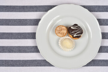Three small, diverse miniature desserts are served on a white plate centered over a gray and white horizontally striped tablecloth