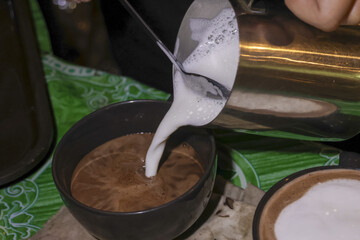 Pouring frothy steamed milk into hot chocolate drink, comforting and warm beverage in rustic cup. This cozy close up shows delicious preparation of sweet coffee treat