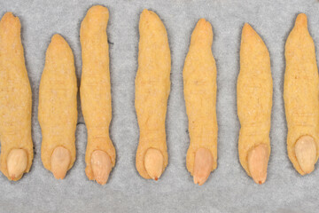 An overhead shot of numerous freshly baked sugar cookies shaped like spooky witch fingers with almond fingernails, laid out on a baking sheet