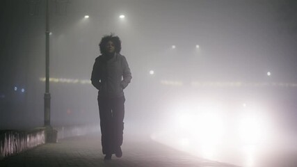A woman walks down a street in the fog. The street is lit up by street lights and car headlights. The scene is dark and mysterious, with the fog adding to the sense of horror - Powered by Adobe
