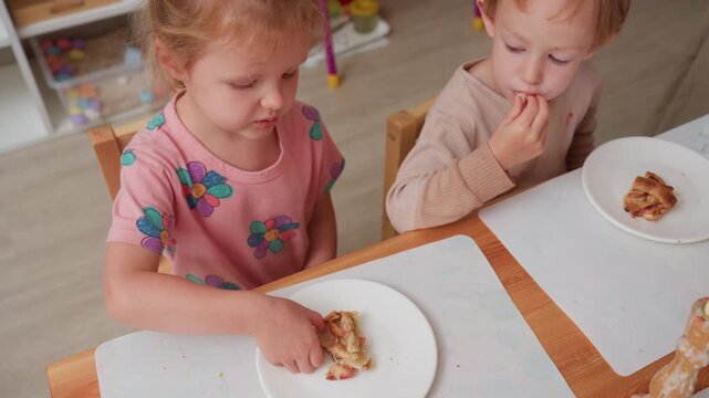 little girl hesitates with dessert, lifts apple pie slice to inspect while boy eats his portion and watches, mixed reactions during preschool snack time at table, focus on picky taste and curiosity