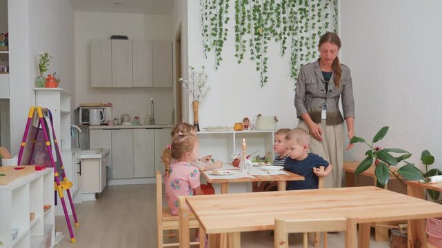 Kindergarten teacher standing beside children sitting at wooden table during lunch, talking to boy in blue shirt inside bright classroom decorated with green plants, educational materials