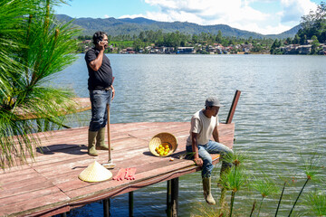 Two Farmers Taking Break on Wooden Dock by Lake one Chatting on His Cell Phone