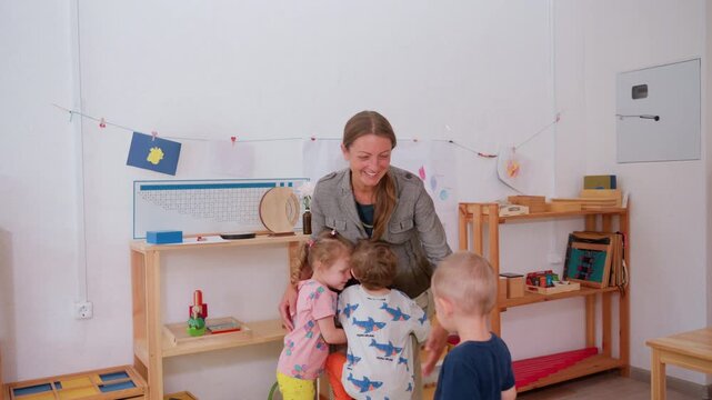 Smiling teacher embraces small children in cheerful classroom surrounded by shelves, drawings, and learning materials, expressing affection, care, and connection during joyful preschool