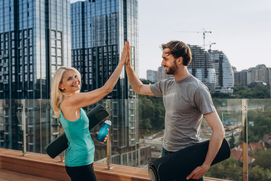 Bearded man and blonde woman give high-five on rooftop with city buildings after yoga session