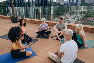 Group of multigenerational people sits in relaxed cross-legged positions on yoga mats on rooftop talking
