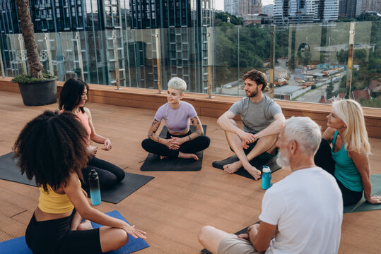 Multigenerational people of different nations sit on rooftop terrace engaged in conversation at yoga break

