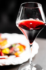 A glass of red wine with a blurry background of a white plate and food. The focus is on the glass, showcasing the rich color of the wine and its elegant shape