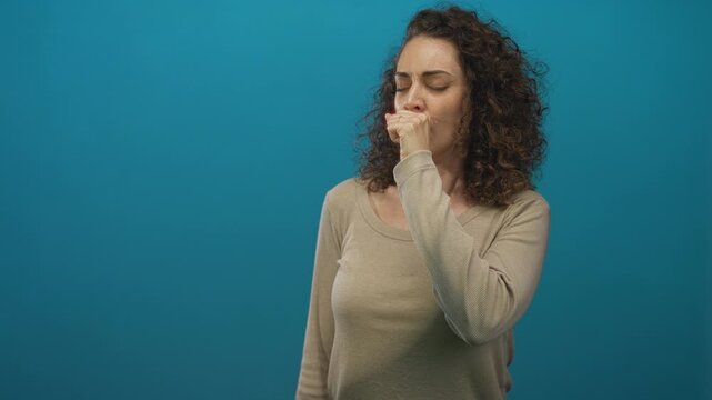 Young woman coughing into her fist in studio with teal backdrop, curly hair and beige long sleeve top; sick recovery.