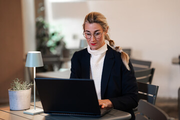 Stylish businesswoman focused on laptop in evening light working indoors with modern elegance and...