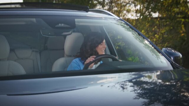 Brunette woman in blue shirt sitting inside car pressing horn with frustration. Female driver reacting to traffic jam annoyed by other motorists. Morning sunlight reflecting on vehicle hood.