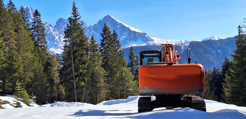 A crawler excavator stands in a snowy forest against the backdrop of Dolomites. Panorama. © Tanya