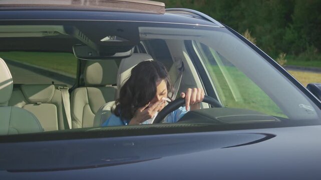 Brunette woman sitting inside car sneezing into tissue paper while holding steering wheel. Female driver with runny nose suffering seasonal allergy. Bright sunlight entering vehicle interior.