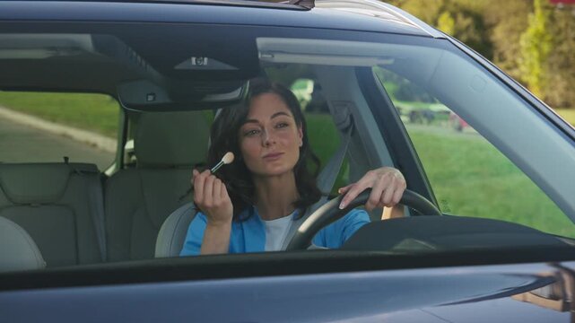 Caucasian woman sitting in car holding makeup brush near face applying powder while driving. Female driver concentrating on appearance before trip. Daytime outside in parked vehicle.