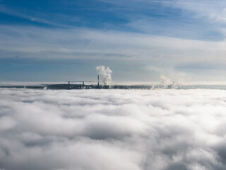Aerial drone view of industrial chimneys emitting steam above thick fog and clouds on a sunny day, power plant and factory landscape. Panorama above clouds in fog in sunny day. Clouds factory