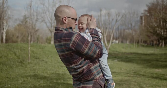 A father holds his baby boy in a park and helps him to walk. The baby is learning to walk with the help of his father. The father also kisses the baby's head.
