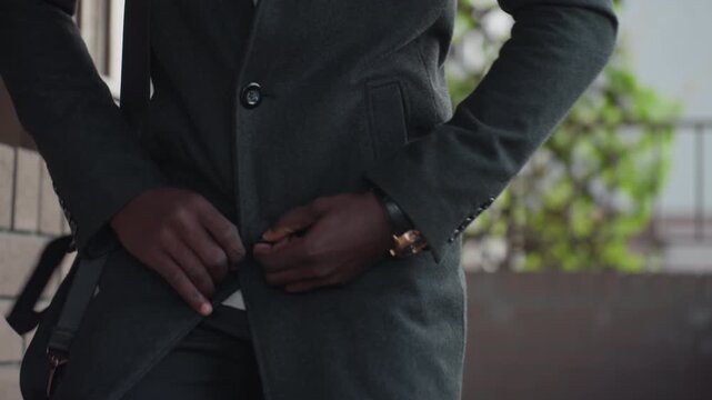 waist view shows hand figure fastening dark wool coat, fingers closing button near buttonhole, wristwatch gleaming, black trousers visible, shoulder bag strap hanging and lattice with fresh leaves