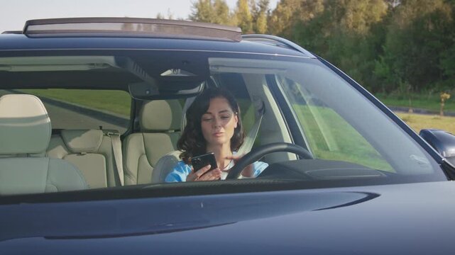 Busy Caucasian woman holding smartphone texting while driving car on sunny road. Female distracted by phone screen instead of watching traffic ahead. Unsafe behavior inside moving vehicle.
