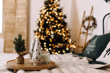 Minimalist holiday home interior with ceramic Christmas trees, small pine, and candle on wooden tray. Blurred Christmas tree lights in the background create a warm festive atmosphere.