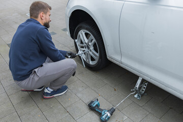 Man changing car tire using electric drill and jack on city sidewalk. DIY auto repair, maintenance, seasonal tire change, mechanical work, vehicle service, urban life.