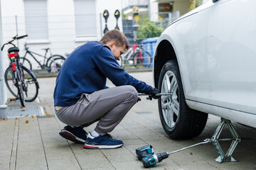 Man changing car tire using electric drill and jack on city sidewalk. DIY auto repair, maintenance, seasonal tire change, mechanical work, vehicle service, urban life.