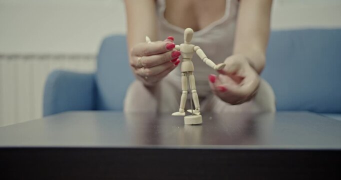 A close-up shot captures a woman's hands with red nail polish posing a small wooden artist's mannequin on a table, highlighting the articulated movements and accompanying sounds.