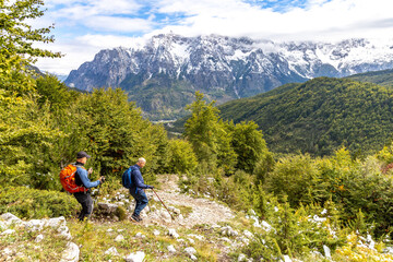 Two men hiking along a rocky mountain trail surrounded by forest, with distant mountains visible in the background on a clear day.