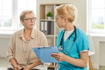 Hospital consultation for senior patient. Young nurse with stethoscope talking to listening serious...