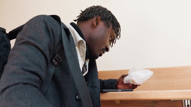 Man with dreadlock bends one side as he leans on table looking at dough and phone, dressed in dark jacket and shirt with turtle neck, showing quiet focus in soft indoor light with realistic calm tone