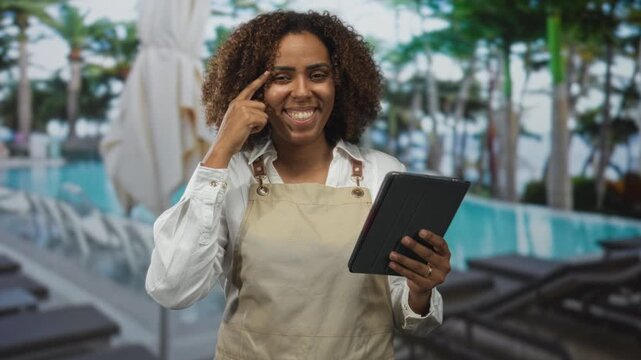 Woman in apron points finger to temple while holding tablet in building pool area, smiling and checking notes on device; confidence hospitality.