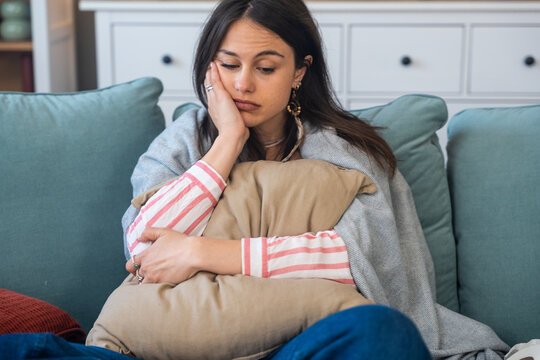 Young woman at home hugging pillow on the sofa feeling unwell with abdominal discomfort possible menstrual cramps stomach ache food poisoning or digestive upset resting under blanket, seeking relief