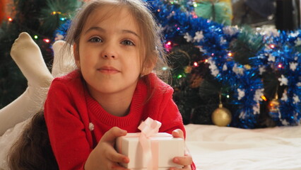 small child baby  girl in a Christmas outfit with a hood near a New Year tree decorated with light...