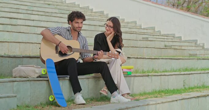 Indian young adult gen z female male man hold guitar play new lyric music sing song have fun joy stairway place. Happy relax teen girl boy guy sitting on stair enjoy good mood spend break day time