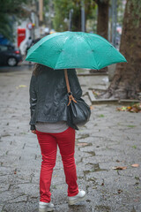 Back view of woman with green umbrella walking on wet pavement in red pants and leather jacket. Urban lifestyle, rainy day, urban solitude