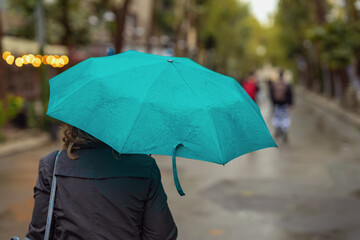 Woman back view with green umbrella walking on rainy city street surrounded by soft bokeh lights and blurred background