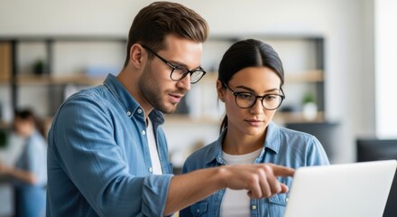 Two colleagues, a man and a woman, wearing glasses and denim shirts, collaborate on a laptop in a modern office setting, pointing and discussing