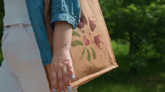 close up of lady walking outdoors carrying embroidered eco bag with colorful floral pattern in sunlight along green bush path, wearing blue shirt and white trousers, symbolizing summer freedom