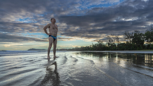 A man in swimming trunks stands on a sandy beach in the surf at dawn. Hands on waist, looking into the distance. The waves spread over the wet sand. The clouds in the sky are highlighted in gold, pink