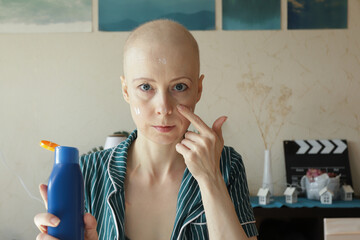 Bald woman in striped pajamas applies sunscreen at home after cancer therapy, showing daily skincare and protection routine for sensitive skin