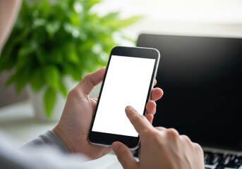 Person holding a smartphone with a blank white screen in front of a laptop and a green plant, closeup shot
